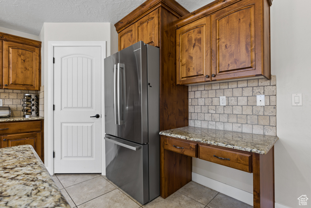 Kitchen featuring freestanding refrigerator, light tile patterned floors, tasteful backsplash, light stone counters, and brown cabinets
