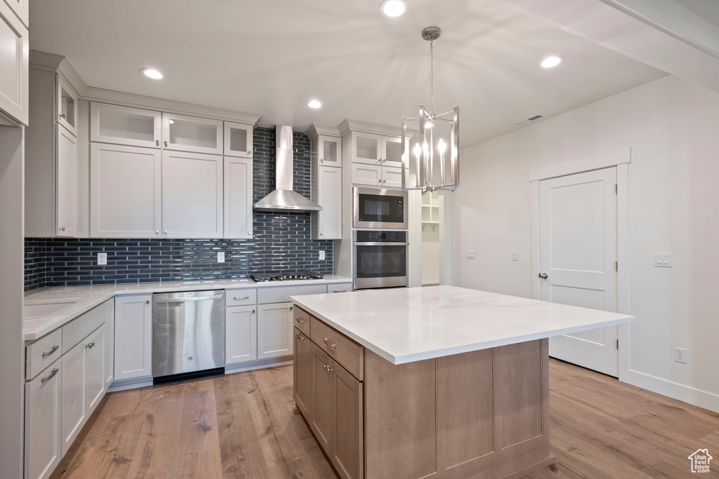 Kitchen featuring appliances with stainless steel finishes, wall chimney range hood, light wood-style flooring, tasteful backsplash, and light countertops