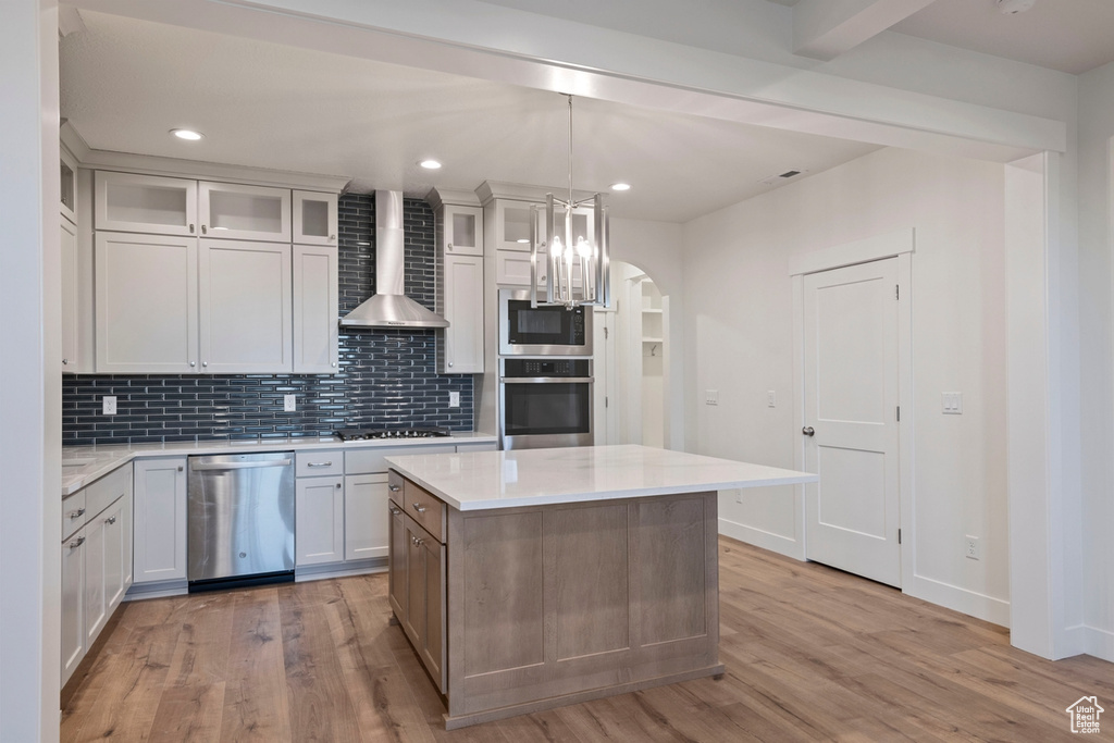 Kitchen with appliances with stainless steel finishes, wall chimney range hood, light wood-type flooring, light countertops, and tasteful backsplash