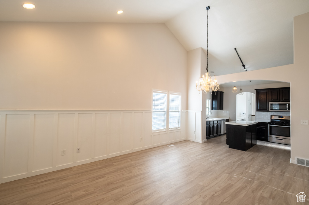 Unfurnished living room with a chandelier, high vaulted ceiling, a decorative wall, a wainscoted wall, and light wood finished floors