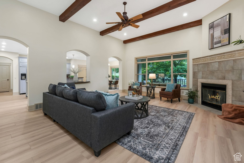 Living room with ceiling fan, light wood-type flooring, beamed ceiling, a tiled fireplace, and arched walkways
