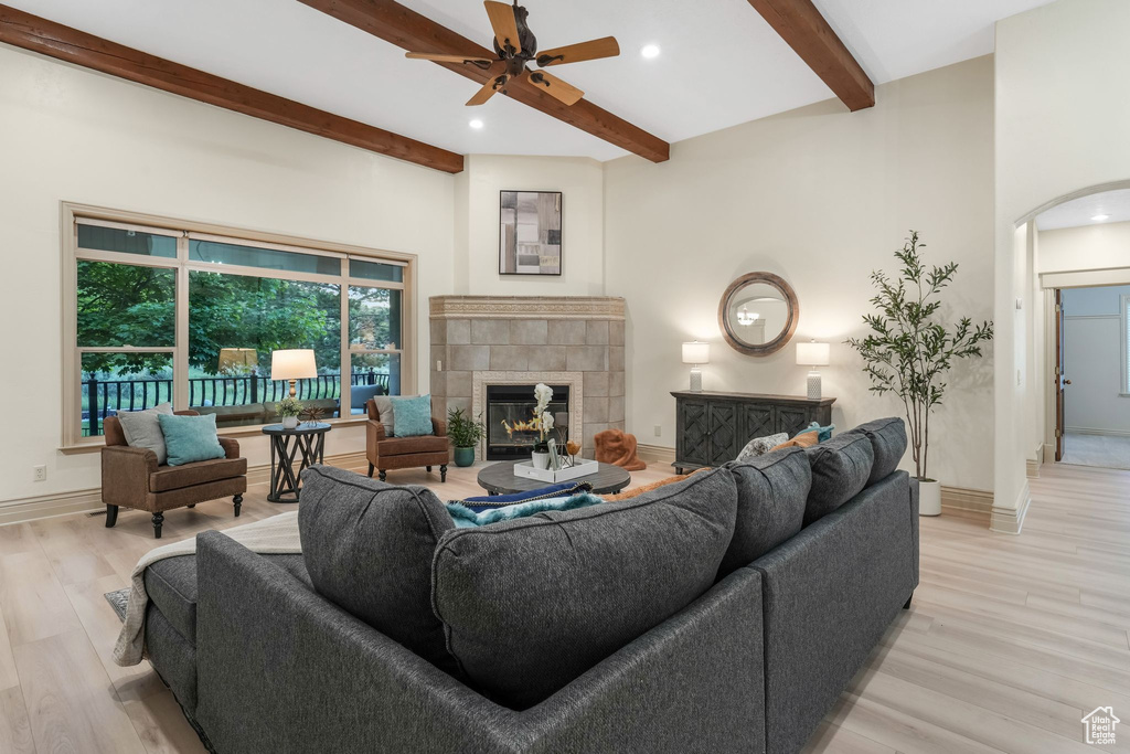Living room featuring arched walkways, beamed ceiling, a tile fireplace, light wood-style floors, and a ceiling fan