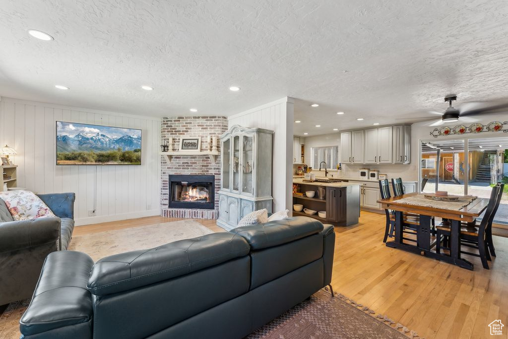 Living room featuring a textured ceiling, light wood-type flooring, a brick fireplace, recessed lighting, and ceiling fan