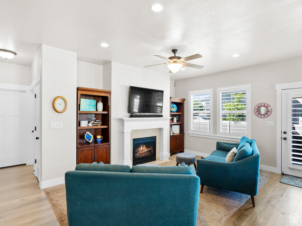 Living room with light wood finished floors, a warm lit fireplace, a ceiling fan, baseboards, and recessed lighting