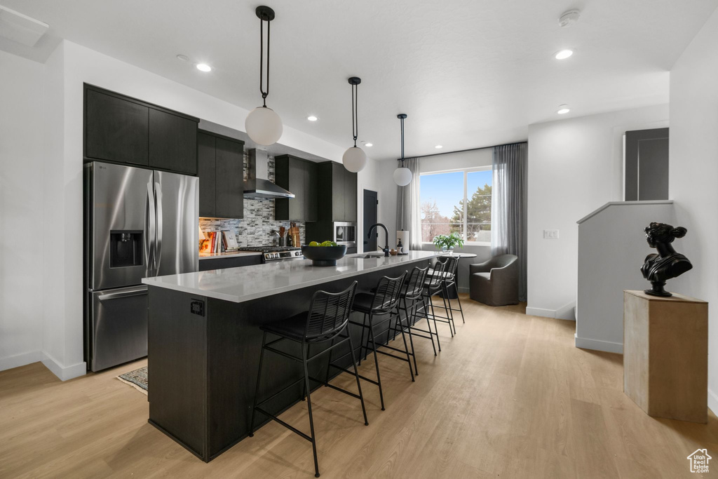 Kitchen featuring appliances with stainless steel finishes, wall chimney range hood, dark cabinets, a large island, and a kitchen breakfast bar