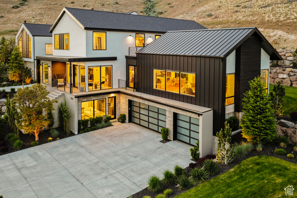 Rear view of house featuring a standing seam roof, concrete driveway, a garage, metal roof, and a balcony
