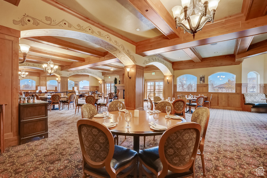 Carpeted dining room featuring a chandelier, beamed ceiling, arched walkways, a wainscoted wall, and decorative columns