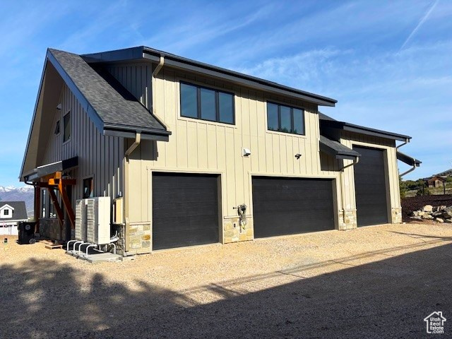 View of side of home featuring stone siding, a garage, dirt driveway, roof with shingles, and board and batten siding