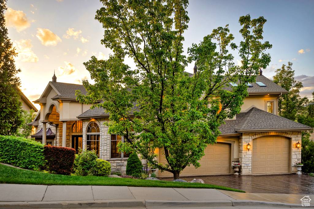 French country home with stone siding, roof with shingles, driveway, and stucco siding