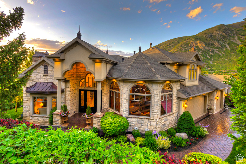 French country style house with stone siding, a garage, a mountain view, a shingled roof, and driveway