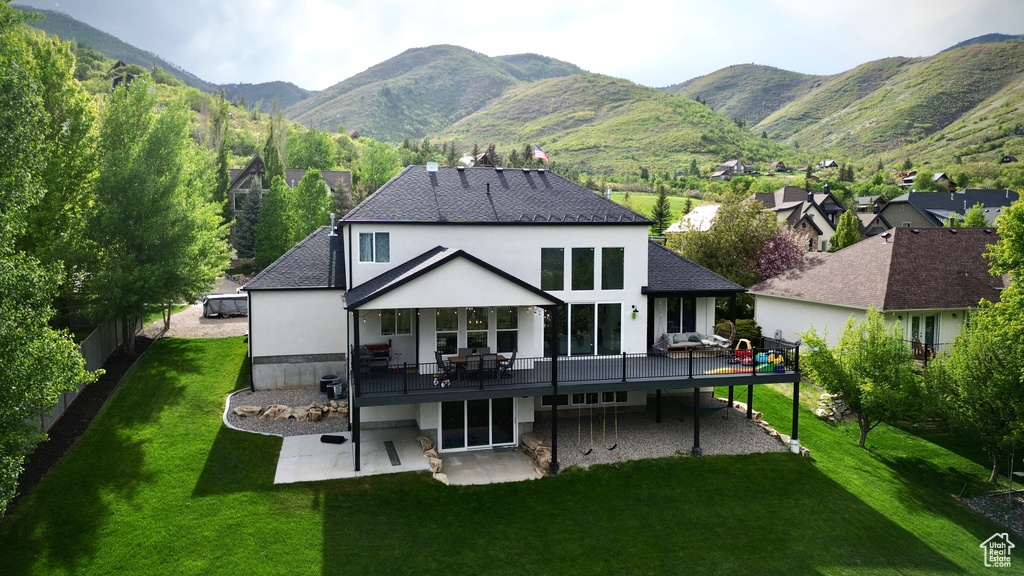 Rear view of house with a patio area, a deck with mountain view, a lawn, a shingled roof, and stucco siding
