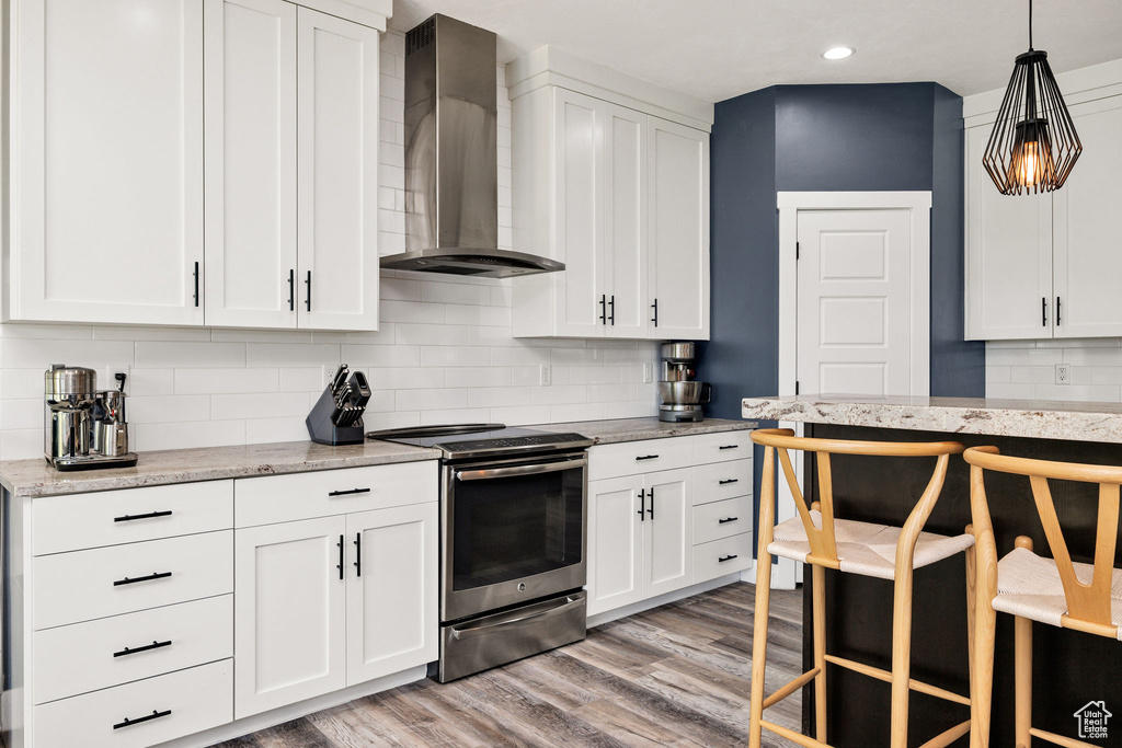 Kitchen featuring stainless steel electric range oven, wall chimney range hood, light wood-type flooring, decorative backsplash, and white cabinetry