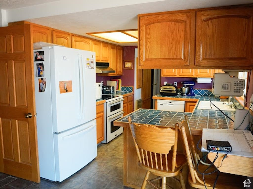Kitchen featuring white appliances, tile counters, under cabinet range hood, and a peninsula