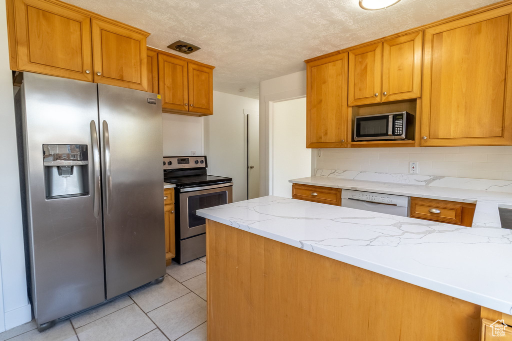 Kitchen with appliances with stainless steel finishes, light stone counters, light tile patterned floors, brown cabinetry, and a textured ceiling