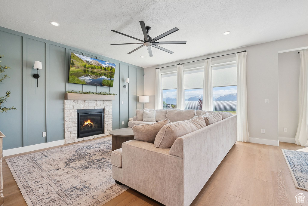 Living area with a ceiling fan, a stone fireplace, light wood finished floors, recessed lighting, and a textured ceiling