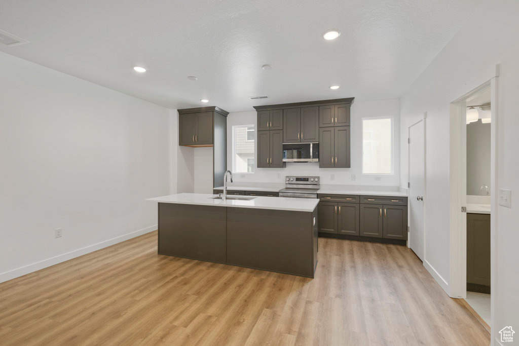Kitchen with stainless steel appliances, a sink, light wood-style floors, light countertops, and an island with sink