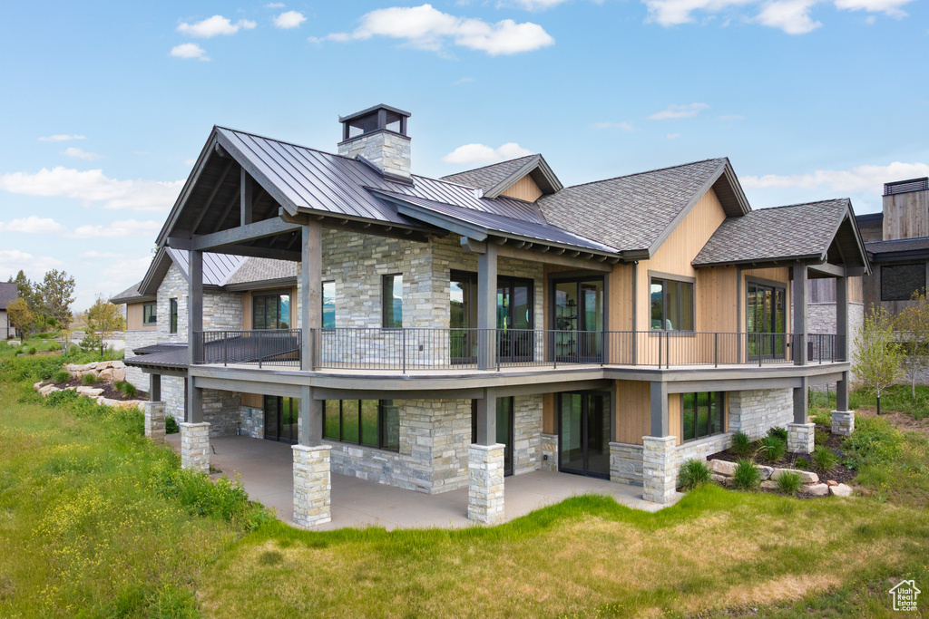 Back of house featuring stone siding, a balcony, a standing seam roof, a chimney, and a patio