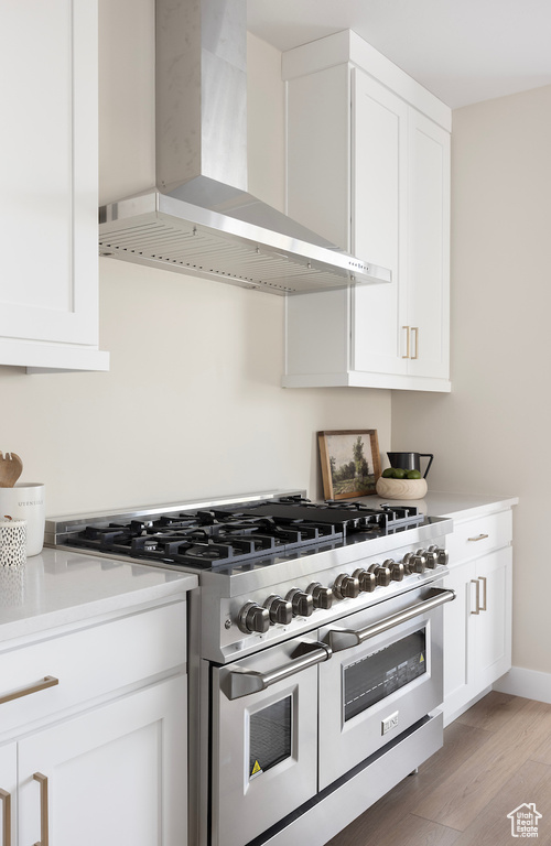 Kitchen with wall chimney range hood, range with two ovens, white cabinetry, light wood-style floors, and light countertops