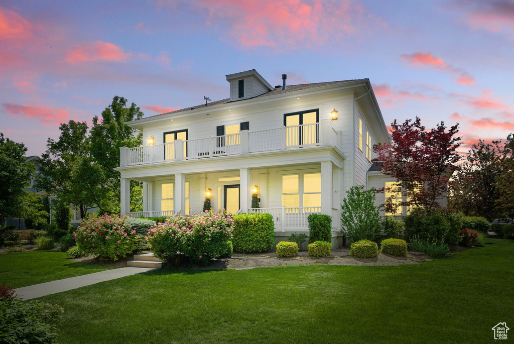 Back of house at dusk featuring a balcony, a porch, and a lawn