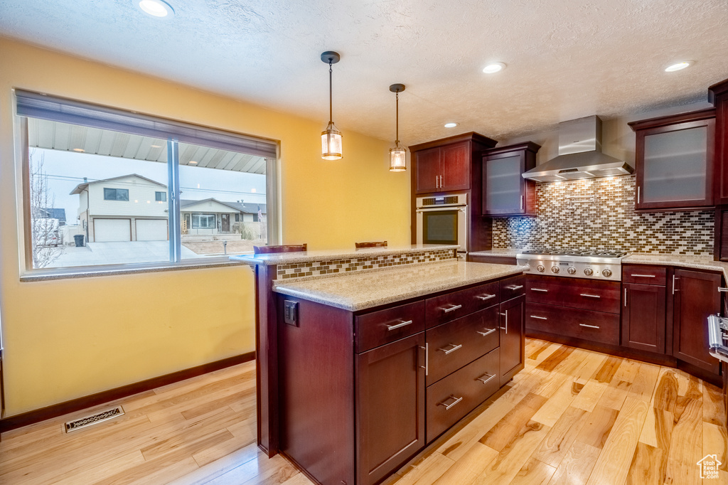 Kitchen featuring wall chimney exhaust hood, appliances with stainless steel finishes, light wood-type flooring, backsplash, and decorative light fixtures