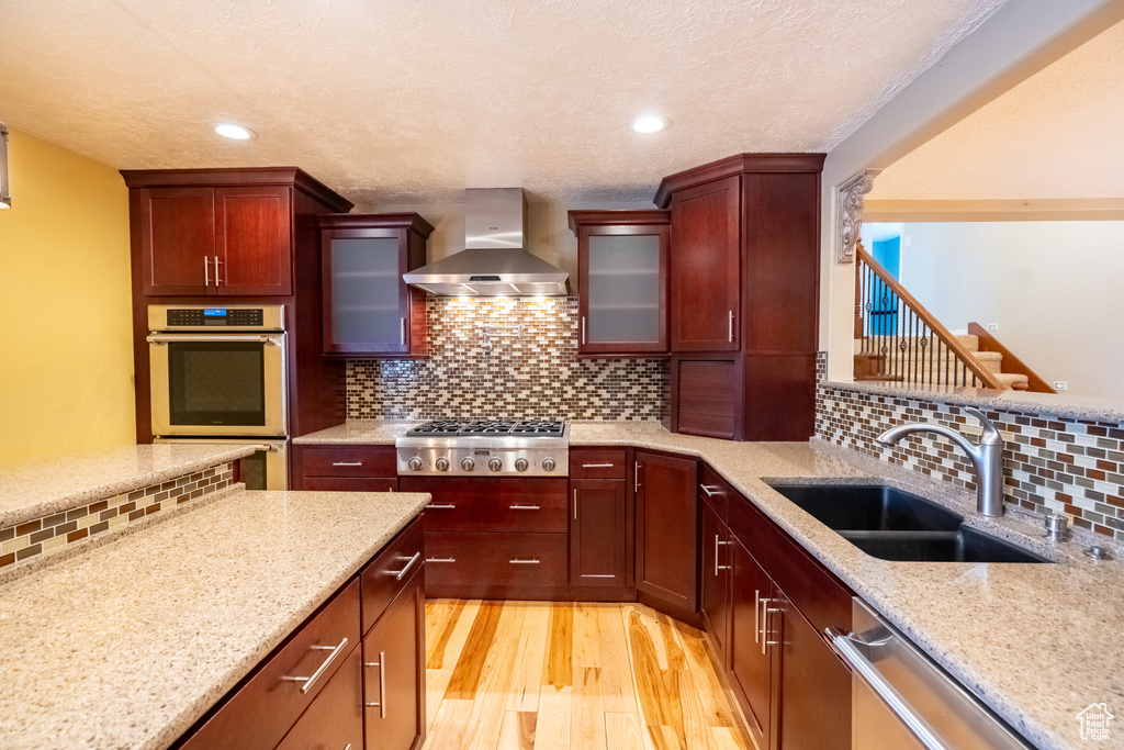 Kitchen with wall chimney exhaust hood, a sink, appliances with stainless steel finishes, light wood-style flooring, and decorative backsplash