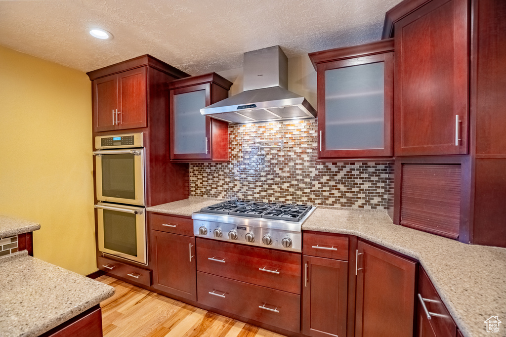 Kitchen with wall chimney range hood, appliances with stainless steel finishes, light wood-type flooring, decorative backsplash, and a textured ceiling