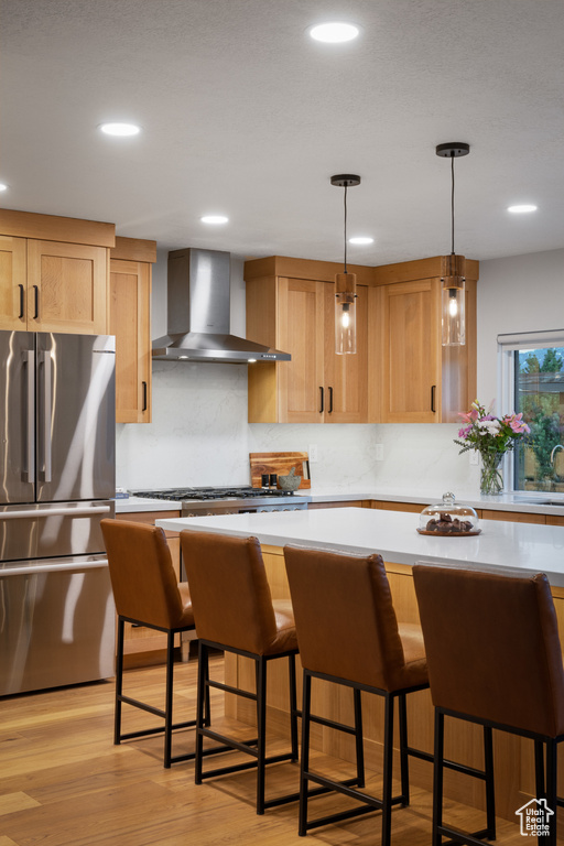 Kitchen with wall chimney range hood, appliances with stainless steel finishes, light countertops, light brown cabinetry, and recessed lighting