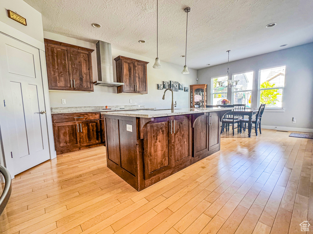 Kitchen featuring wall chimney exhaust hood, light wood-type flooring, an island with sink, a chandelier, and a textured ceiling