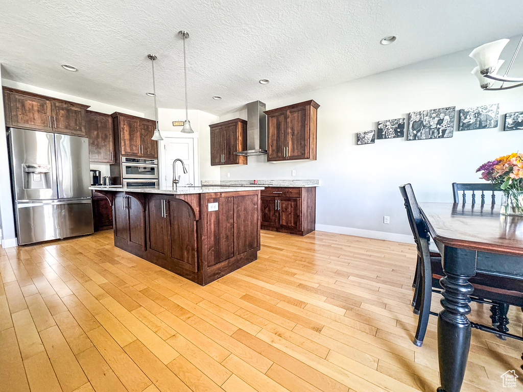 Kitchen featuring stainless steel appliances, wall chimney exhaust hood, a kitchen breakfast bar, light wood-style flooring, and light countertops
