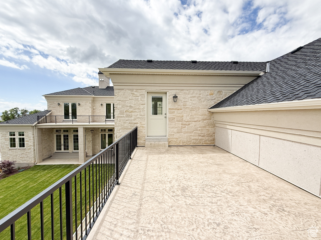 Back of house with a balcony, stone siding, a yard, roof with shingles, and a patio area
