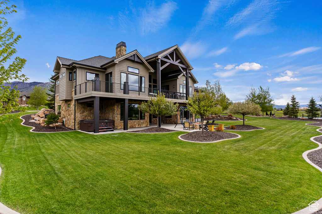 Back of house with a hot tub, a patio area, stone siding, a chimney, and a lawn
