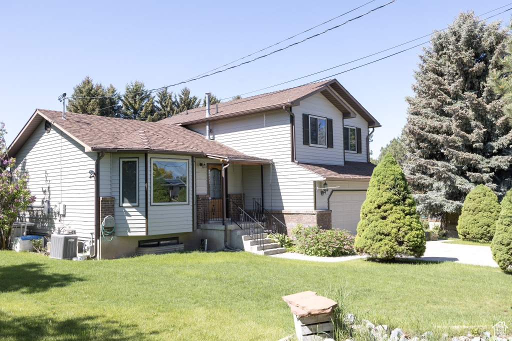 View of front facade with a garage, a front yard, roof with shingles, and cooling unit
