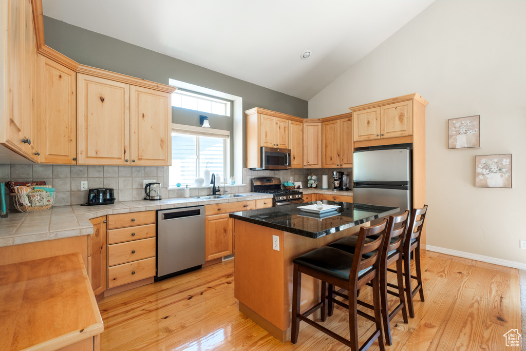 Kitchen with stainless steel appliances, light brown cabinetry, a breakfast bar area, decorative backsplash, and light wood-style flooring