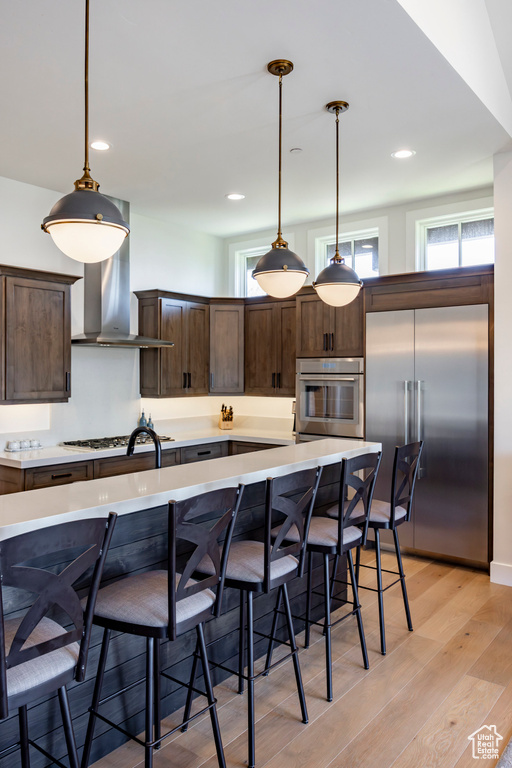 Kitchen with stainless steel appliances, dark brown cabinetry, wall chimney range hood, light countertops, and light wood-type flooring
