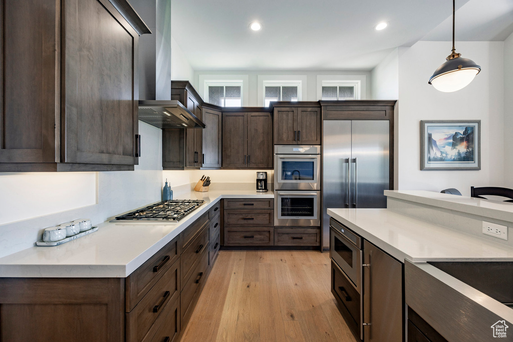 Kitchen with built in appliances, dark brown cabinetry, light wood-type flooring, wall chimney range hood, and light countertops