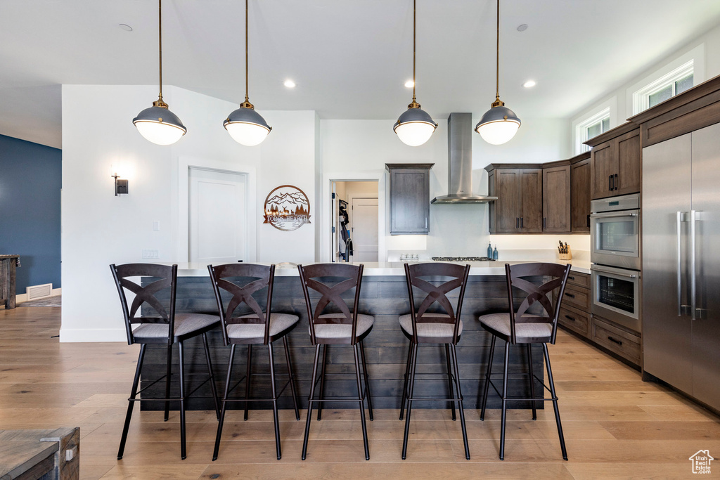 Kitchen featuring wall chimney range hood, stainless steel appliances, dark brown cabinets, light countertops, and a kitchen breakfast bar