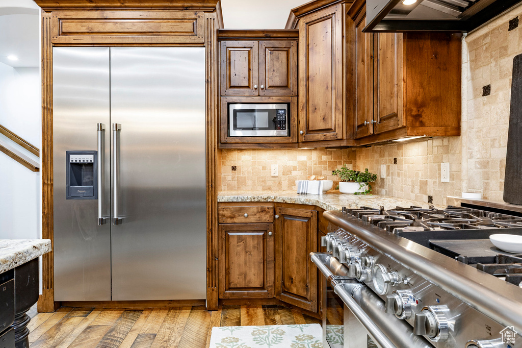 Kitchen featuring built in appliances, light stone counters, backsplash, and under cabinet range hood