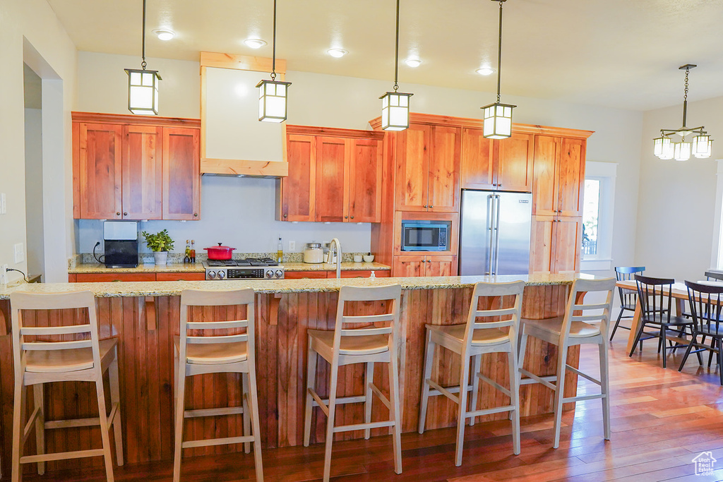 Kitchen with stainless steel appliances, light wood-style flooring, wall chimney exhaust hood, decorative light fixtures, and healthy amount of natural light