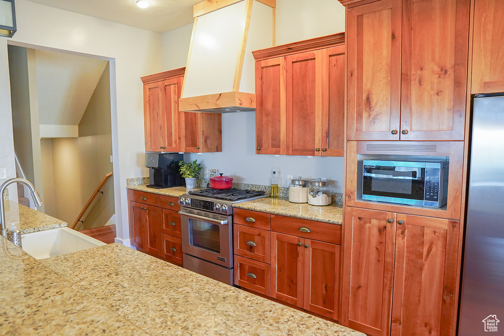 Kitchen with stainless steel appliances, premium range hood, a sink, light stone counters, and brown cabinetry