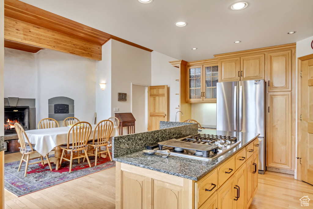 Kitchen featuring stainless steel appliances, light wood finished floors, a warm lit fireplace, light brown cabinets, and dark stone countertops