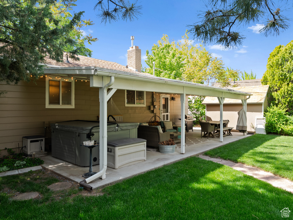 Back of house with a hot tub, a patio area, a chimney, a lawn, and roof with shingles