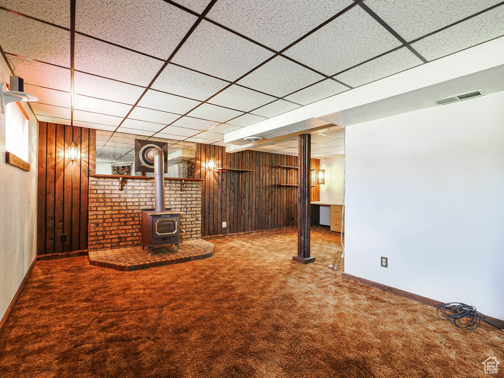 Unfurnished living room with a drop ceiling, carpet flooring, a wood stove, wood walls, and baseboards