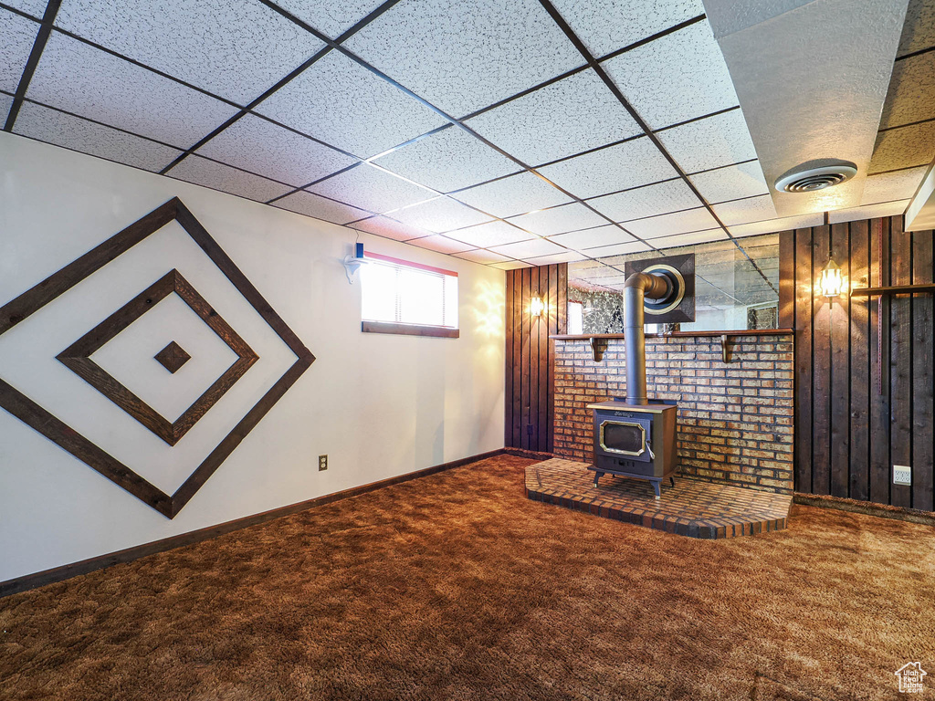 Unfurnished living room featuring carpet flooring, a wood stove, a paneled ceiling, baseboards, and wood walls