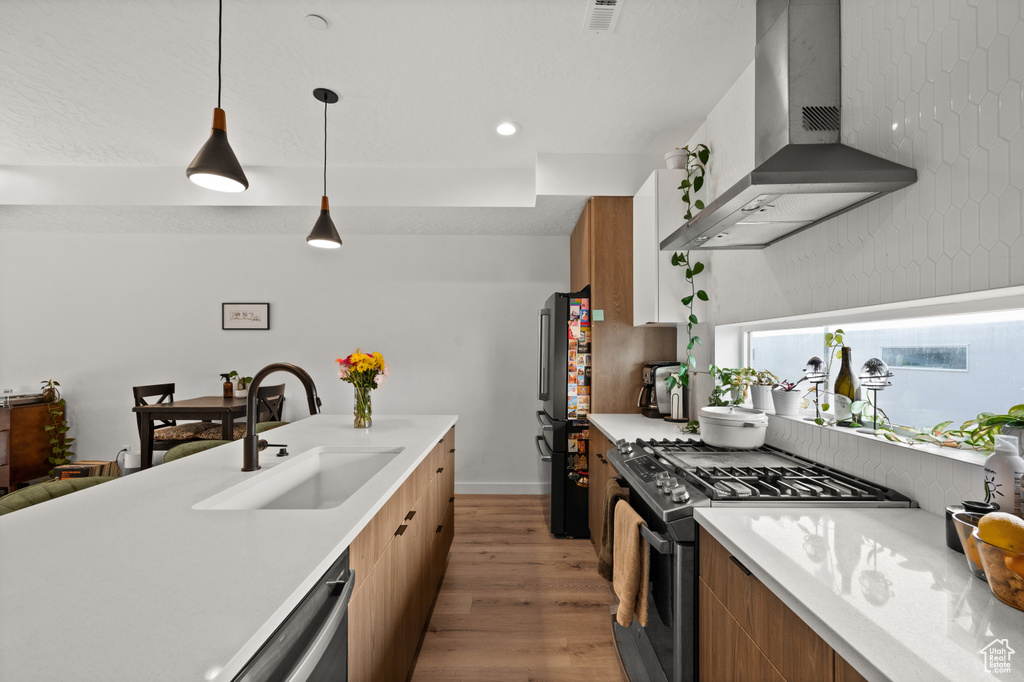 Kitchen with stainless steel appliances, a sink, wall chimney exhaust hood, light wood-type flooring, and brown cabinets