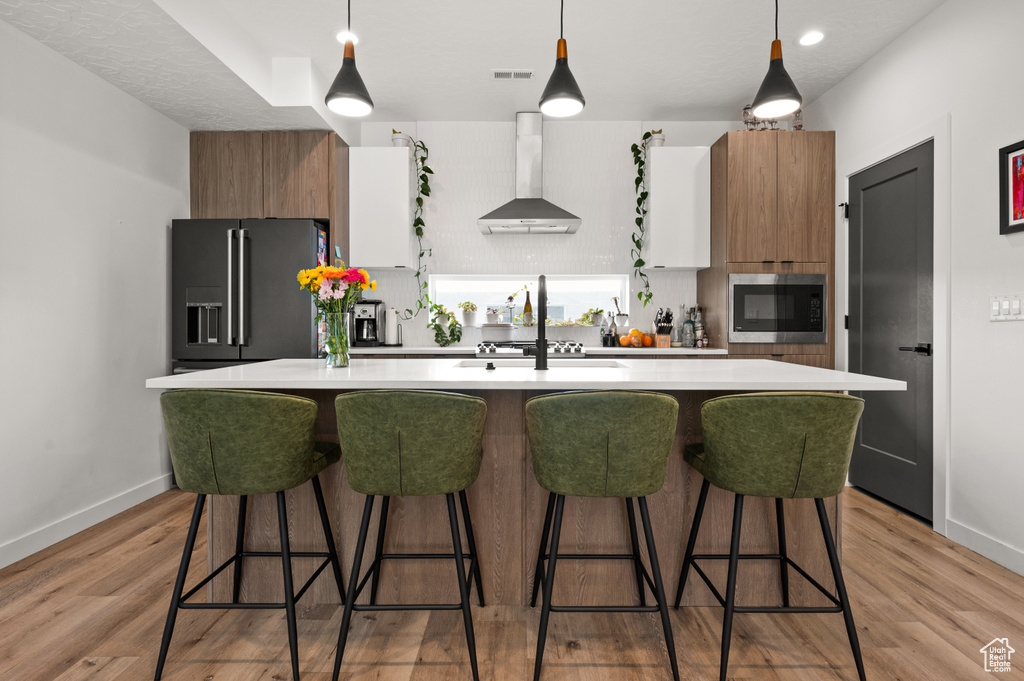 Kitchen with wall chimney exhaust hood, high quality fridge, stainless steel microwave, light countertops, and an island with sink