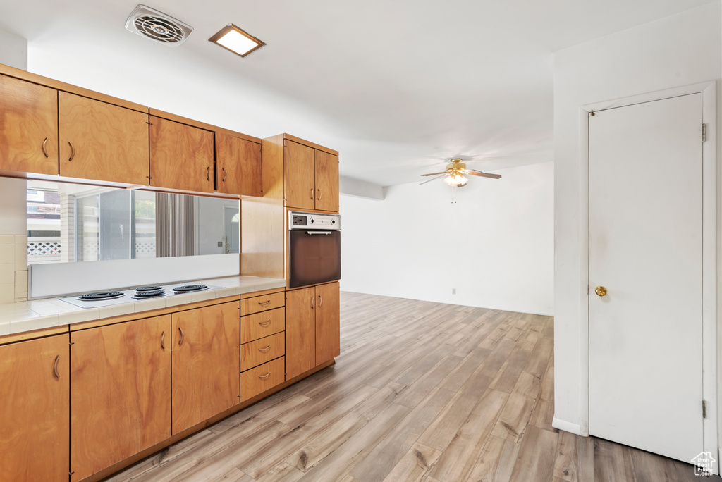 Kitchen featuring wall oven, white electric stovetop, light wood finished floors, ceiling fan, and tile counters