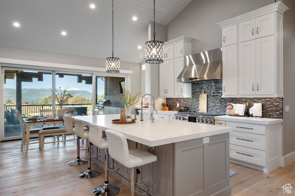 Kitchen featuring wall chimney range hood, backsplash, light wood finished floors, light countertops, and an island with sink
