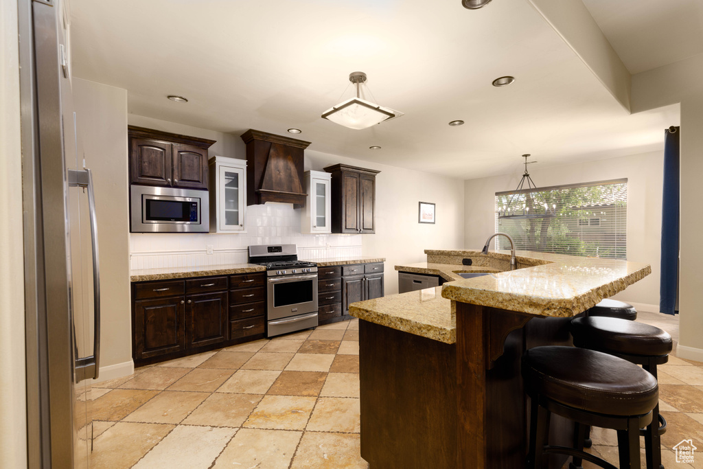 Kitchen with appliances with stainless steel finishes, custom exhaust hood, a sink, dark brown cabinetry, and a kitchen breakfast bar