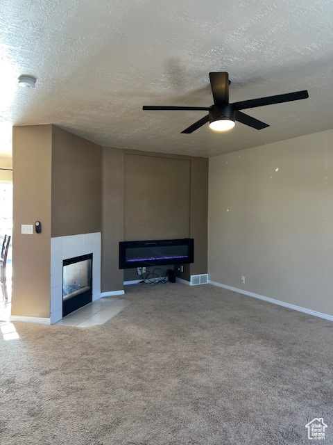 Unfurnished living room featuring a tiled fireplace, carpet flooring, a textured ceiling, a ceiling fan, and baseboards
