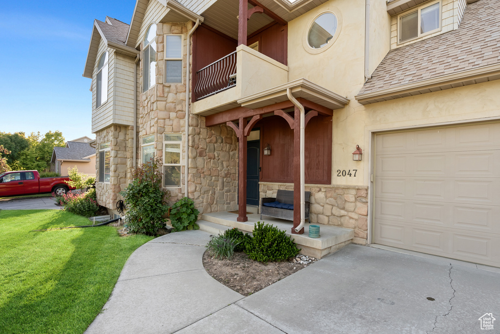 Entrance to property featuring a balcony, stone siding, a lawn, and roof with shingles